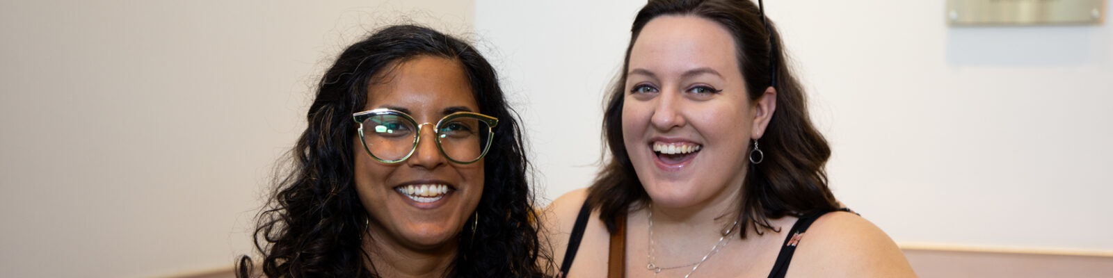 Two people standing next to each other and smiling at an Alumni event. They are standing near a table of swag for attendees. One is carrying a purple baseball cap, branded for Emerson College alumni.